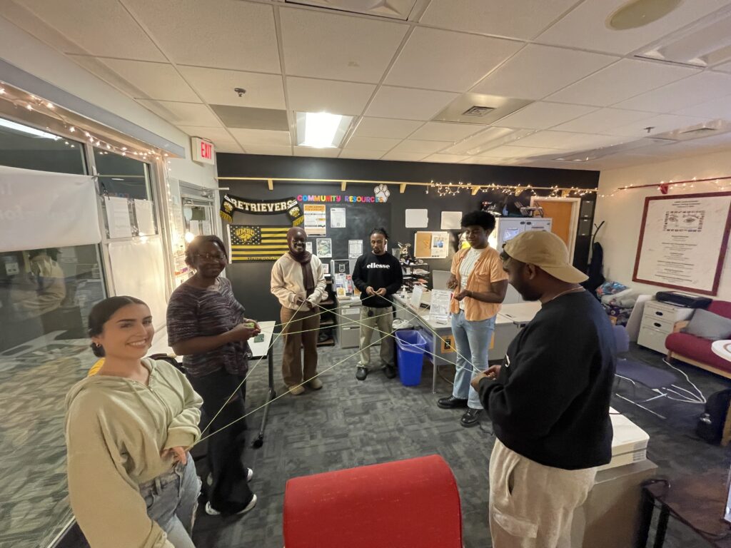 Image of students with diverse background sitting in a dialogue circle engaged in web of connection activity where yarn demonstrates the connection they experienced completing the activity.
